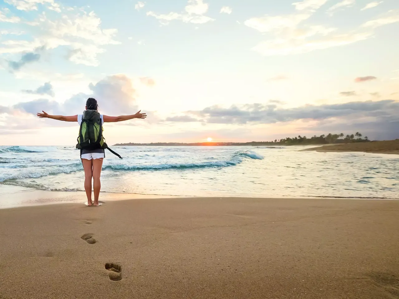 klimaVest: Eine
  Frau mit Rucksack steht am Strand und schaut sich mit offenen Armen das Meer
  und die Sonne an, ihre Fußspuren sind man im Sand.