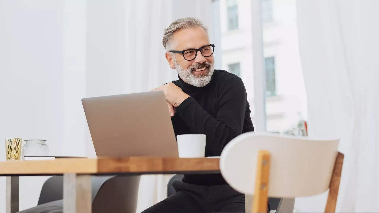 Ein Mann mittleren Alters sitzt in einem schwarzen Rollkragenpullover und einer schwarzen Hose vor einem Laptop, welches auf einem Holztisch platziert ist, und einer Tasse Kaffee in der Hand in einem weißen Zimmer seiner Wohnung und blickt lächelnd nach links; auf dem Tisch steht eine Vase aus Glas und eine goldene Kerze.