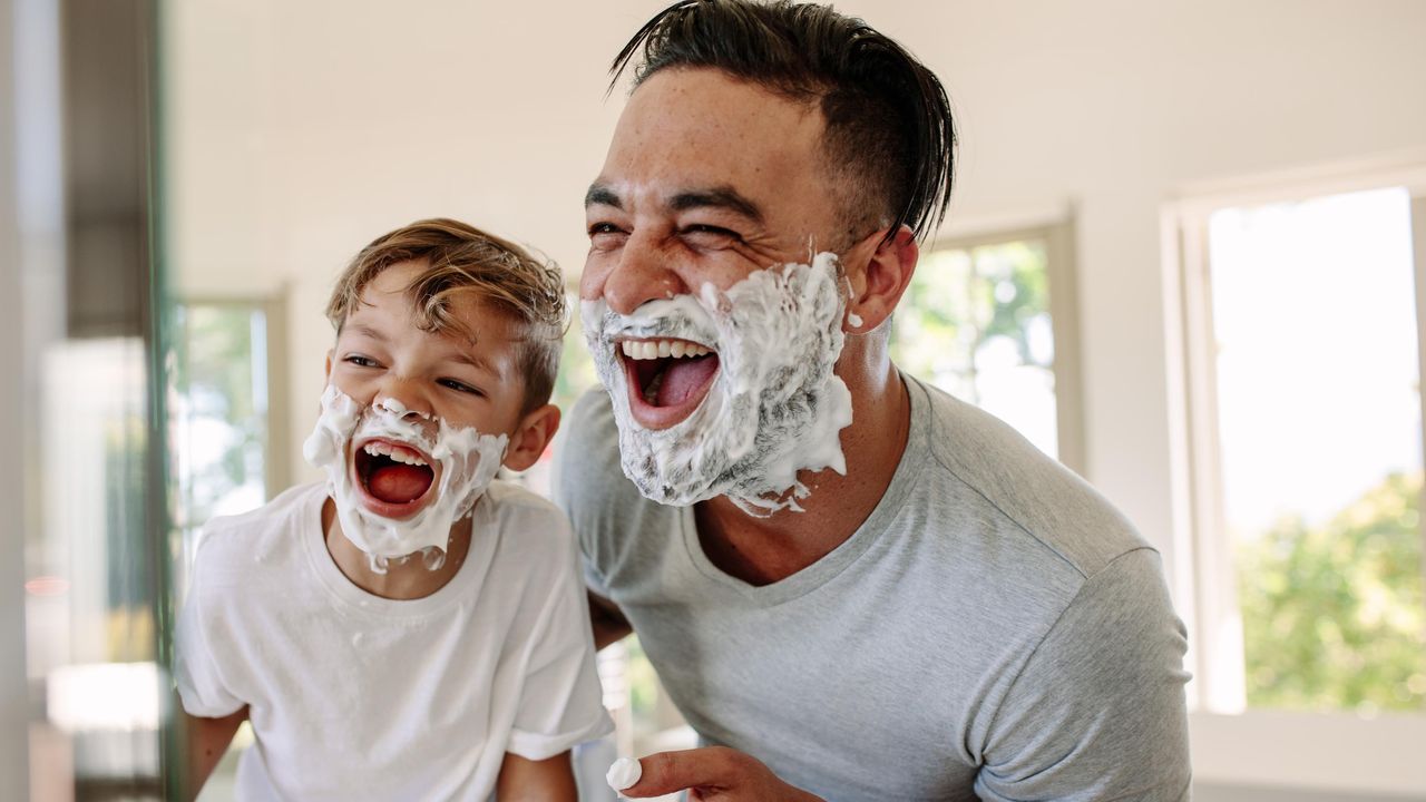Ein lachender Mann zeigt seinem Sohn wie man sich rasiert.