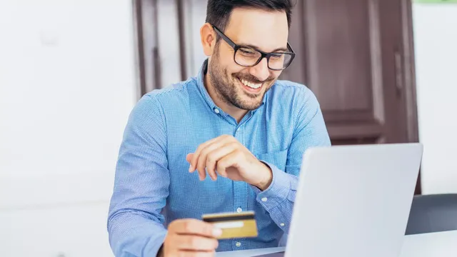 Mann in einem blau-karierten Hemd blickt lächelnd auf seinen Laptop und hat dabei eine Kreditkarte in der Hand.
