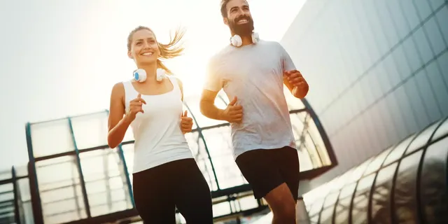 Zwei SportlerInnen mit Kopfhörern  – ein lächelnder Mann und eine lächelnde Frau – rennen draußen vor glasüberzogenen Gebäude.