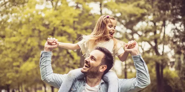 Die blondhaarige Tochter in einem weißen T-Shirt sitzt auf den Schultern ihres Vaters in einem hellblauen Jeanshemd und einem weißen T-Shirt; die beiden befinden sich in einem Park.