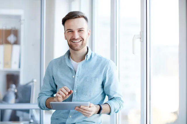 Ein braunhaariger junger Mann in einem weißen T-Shirt und einem hellblauen Jeanshemd hält seinen Tablet in einem sonnigen Büro stehend und lächelt in die Kamera.