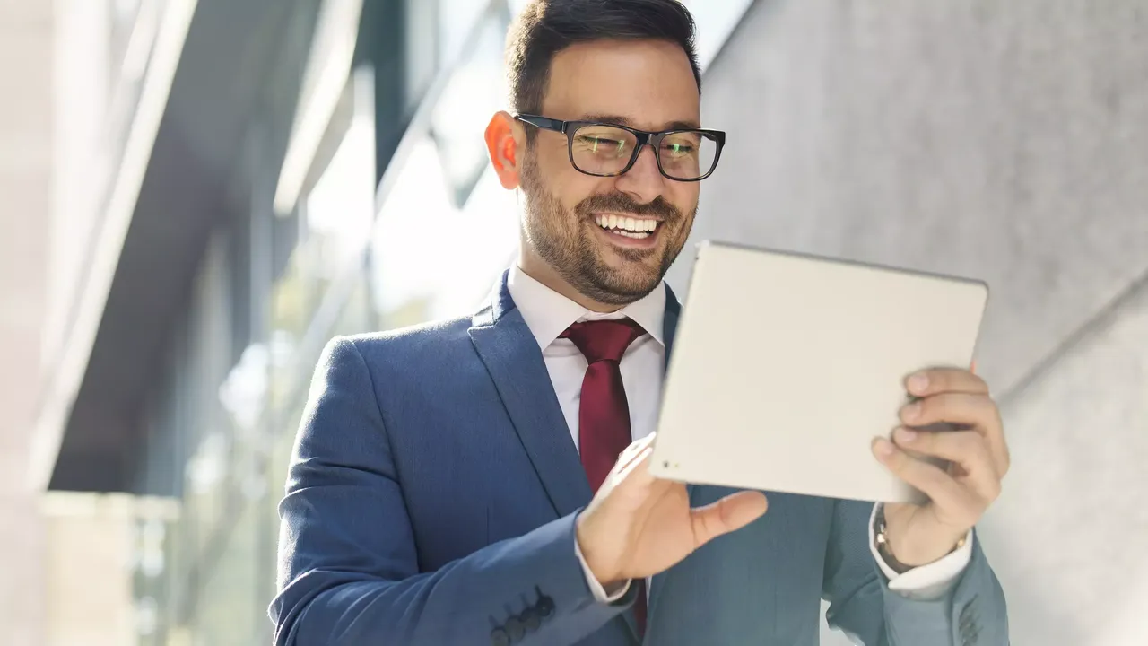 Ein junger dunkelhaariger Mann in schwarzer moderner Brille in einem weißen Hemd, einem dunklen Anzug mit einer roten Krawatte steht draußen vor einem Gebäude und lächelt, auf sein Tablet blickend.