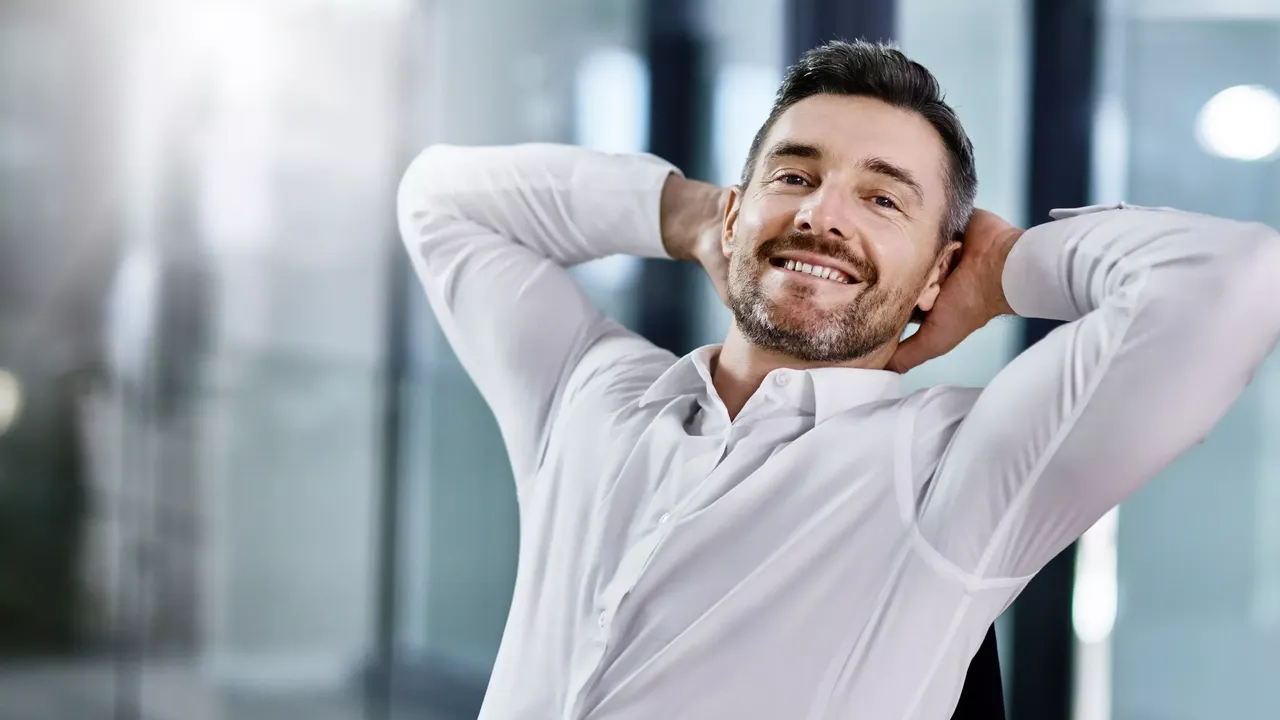 Ein lächelnder Mann mittleren Alters in einem weißen Hemd sitzt im Bureau mit seinen Händen hinter dem Kopf.