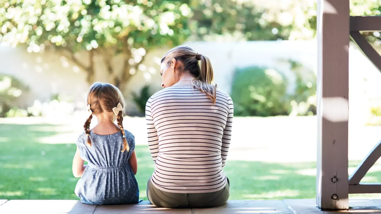 Die Mutter mit hellen Haaren in einem gestreiften T-Shirt mit ihrer kleinen Tochter in einem blauen Kleid und Zöpfen sitzen auf der Terrasse in ihrem Garten.