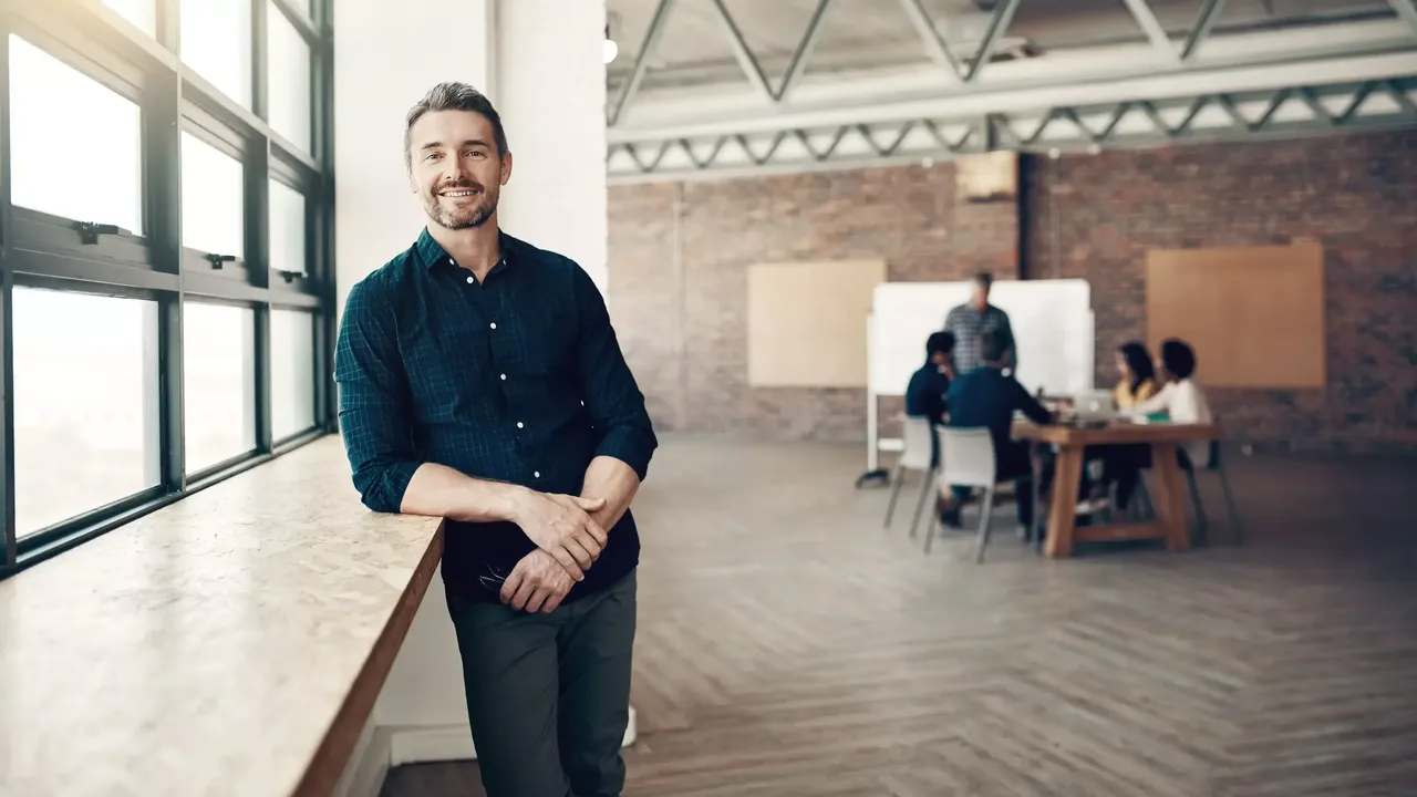 Halb auf das Fensterbrett in einem geräumigen Co-Working Raum mit braunroten Ziegelwänden gelehnt, blickt ein lächelnder bärtiger Mann in einem blauen karierten Hemd direkt in die Kamera; im unscharf aufgenommenen Hintergrund hält vor seinen KollegInnen ein Mann eine Präsentation.