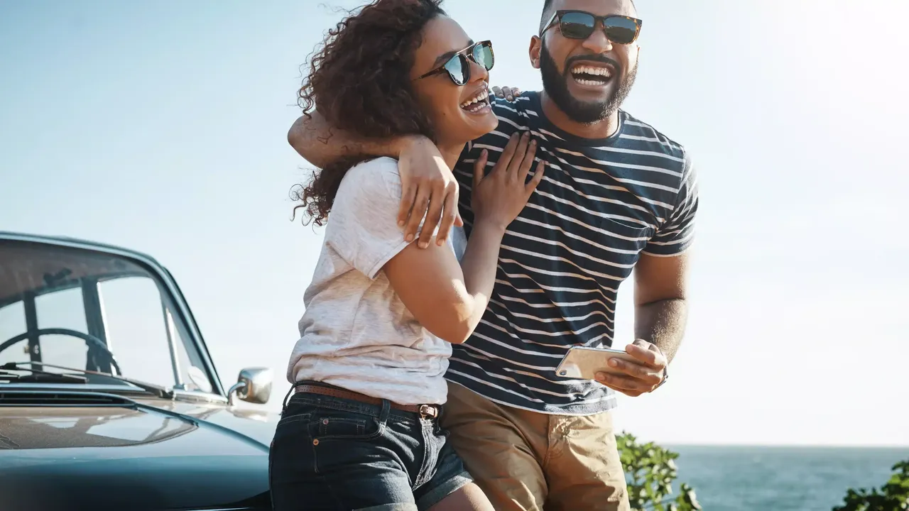 Zwei lächelnde Personen – ein Mann in einem gestreiften T-Shirt und einer Sonnenbrille und eine Frau in einem weißen T-Shirt und einer Sonnenbrille – umarmen sich vor einem Auto stehend; im Hintergrund ist ein Meer zu sehen.