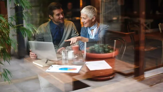 Ein dunkelhaariger Mann in einem dunkelblauen Hemd und einem grauen Cardigan richtet sein Blick auf die Blondine in einem hell-blauen Cardigan, welche auf den Bildschirm mit dem Finger zeigt; die beiden sitzen im Restaurant am Holztisch vor zwei Laptops und dem farbigen Tortendiagramm auf dem Tisch.