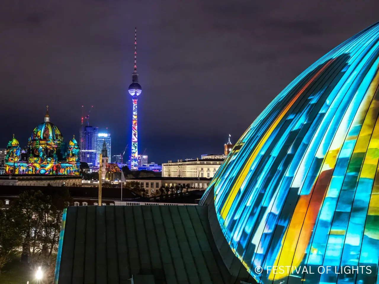 Es wird das Festival of Lights 2025 mit dem illuminierten Alexanderplatz und im Hintergrund die hausInvest Projektentwicklung The Berlinian dargestellt. 