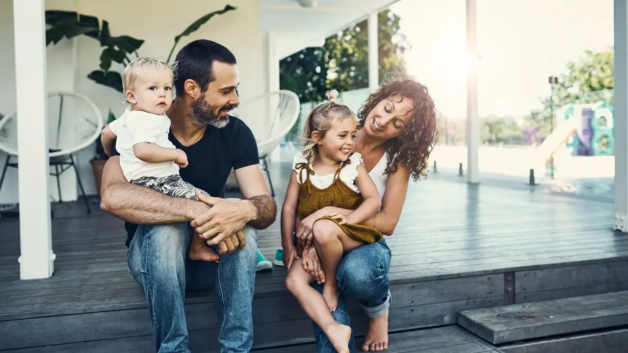 Eine Familie – ein bärtiger Mann in einem schwarzen T-Shirt und Jeans, ein kleiner Junge in einem weißen T-Shirt und Shorts mit einem Pflanzenmuster, ein kleines Mädchen in einem khakifarbigen Kleid und eine Frau in einem weißen T-Shirt und Jeans – sitzen draußen auf der graufarbigen Holzveranda ihres Hauses und lächeln; im Hintergrund sind zwei weiße Stühle und eine Pflanze in einem Topf zu sehen.