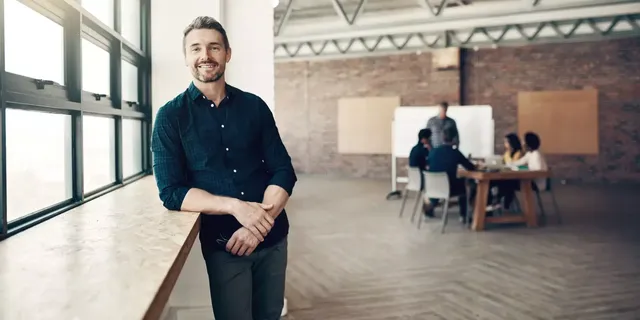 Halb auf das Fensterbrett in einem geräumigen Co-Working Raum mit braunroten Ziegelwänden gelehnt, blickt ein lächelnder bärtiger Mann in einem blauen karierten Hemd direkt in die Kamera; im unscharf aufgenommenen Hintergrund hält vor seinen KollegInnen ein Mann eine Präsentation.