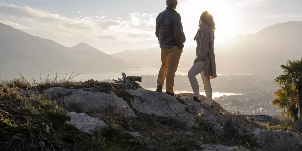 Älteres Paar bei einer Wanderung entlang sonnigen Bergrücken, Sonnenuntergang mit Blick auf eine Stadt mit vielen Immobilien