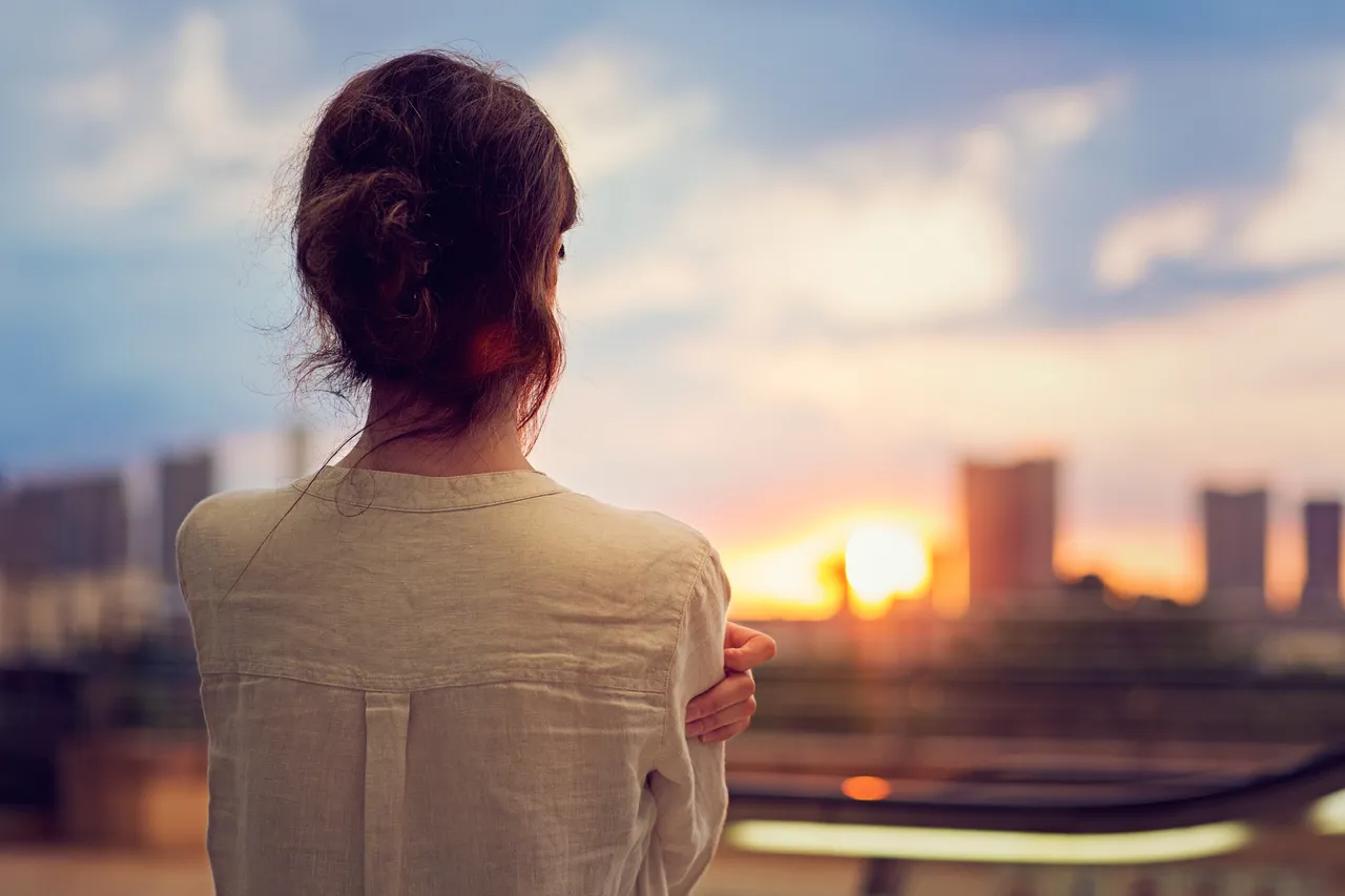 Eine Frau mit einer weißen Bluse und braunen Haaren schaut auf eine Skyline bei Sonnenuntergang.