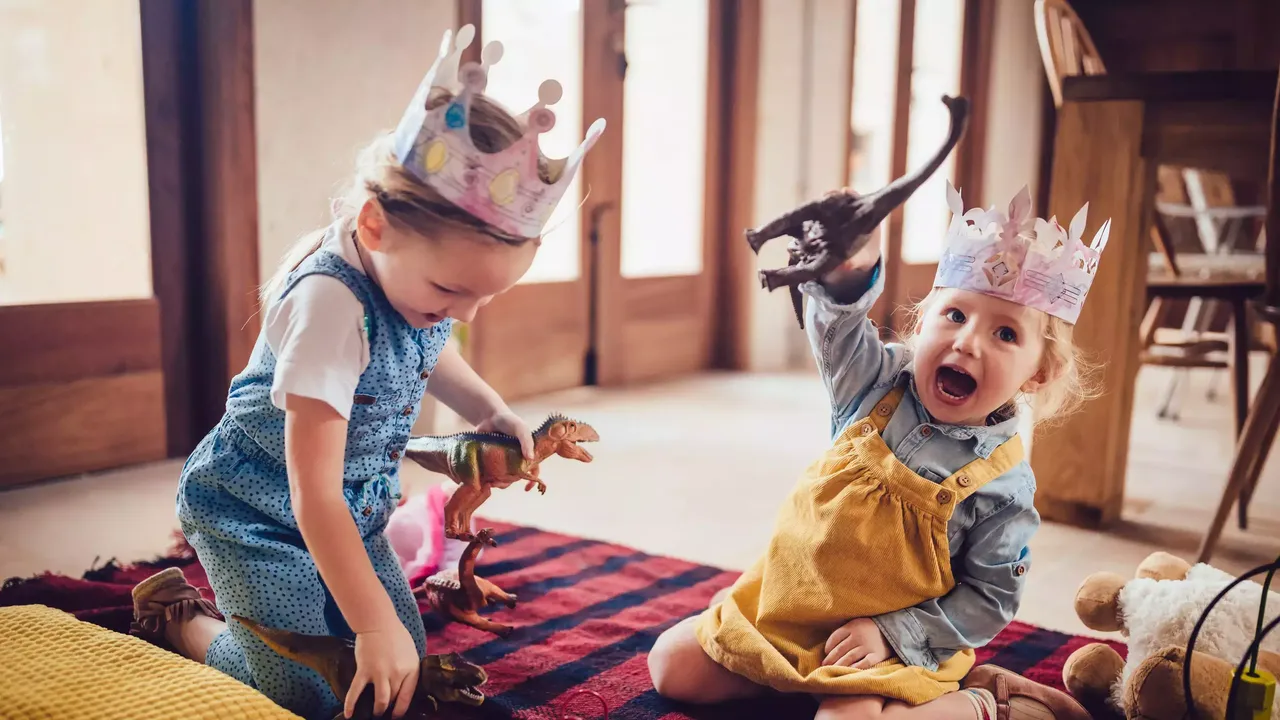 Zwei hellhaarige Mädchen – in einem blauen und einem gelben Kleid – sitzen in pinken Papierkronen auf dem Boden eines Wohnzimmers umgeben von vielen Spielzeugen und spielen mit Dinosaurier-Figuren