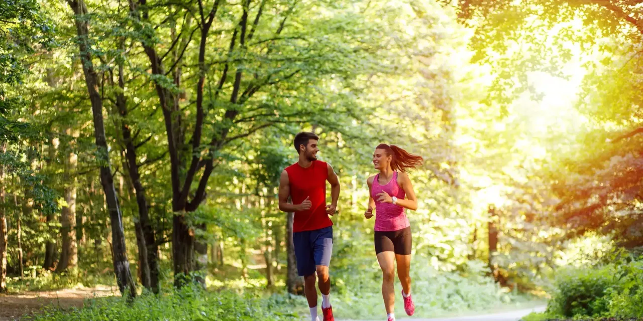 klimaVest: Ein
  Mann und eine Frau joggen auf einer Straße im Park. Im Hintergrund scheint
  die Sonne.