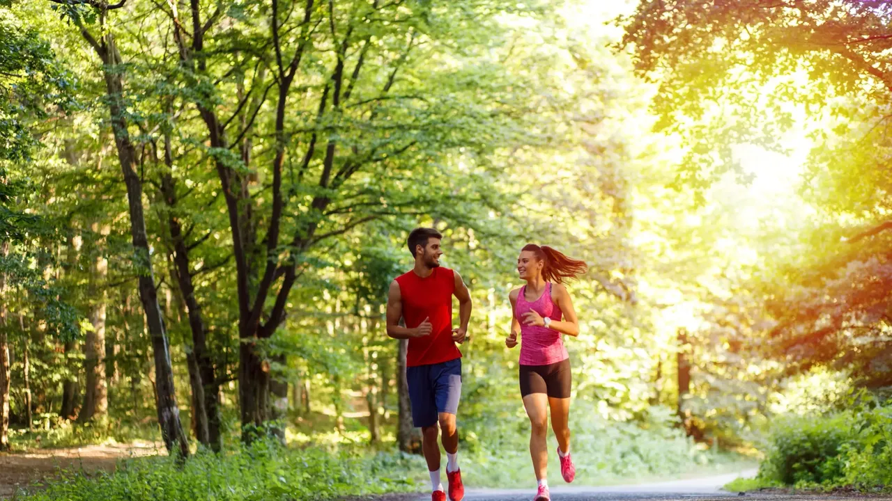 klimaVest: Ein
  Mann und eine Frau joggen auf einer Straße im Park. Im Hintergrund scheint
  die Sonne.