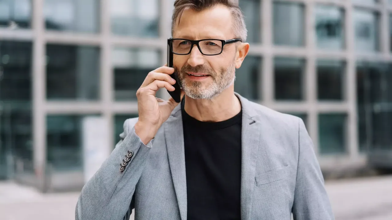 Ein Mann mittleren Alters in schwarzer Brille steht in einem hellen Anzug und einem schwarzen T-Shirt draußen vor seinem Office und telefoniert mit jemanden am Smartphone.