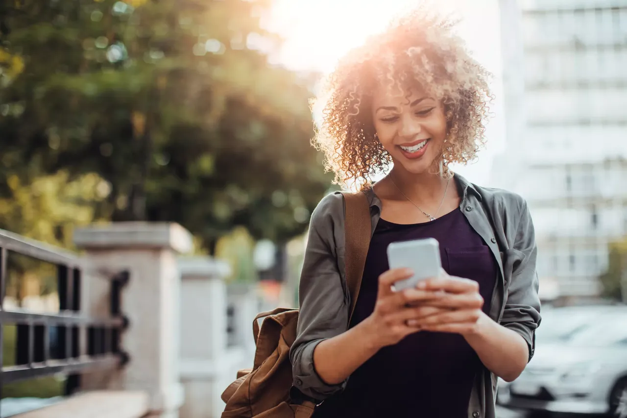 klimaVest: Eine
  Frau mit Handy in der Stadt und Bäumen im Hintergrund.