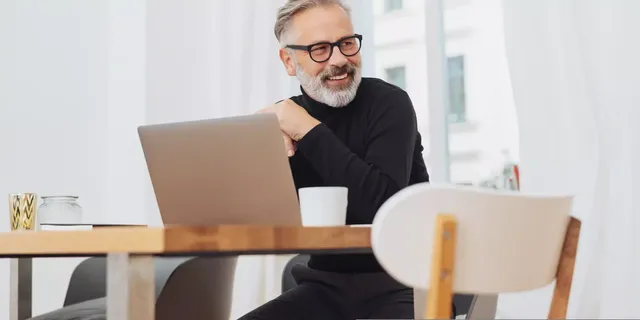 Ein Mann mittleren Alters sitzt in einem schwarzen Rollkragenpullover und einer schwarzen Hose vor einem Laptop, welches auf einem Holztisch platziert ist, und einer Tasse Kaffee in der Hand in einem weißen Zimmer seiner Wohnung und blickt lächelnd nach links; auf dem Tisch steht eine Vase aus Glas und eine goldene Kerze.