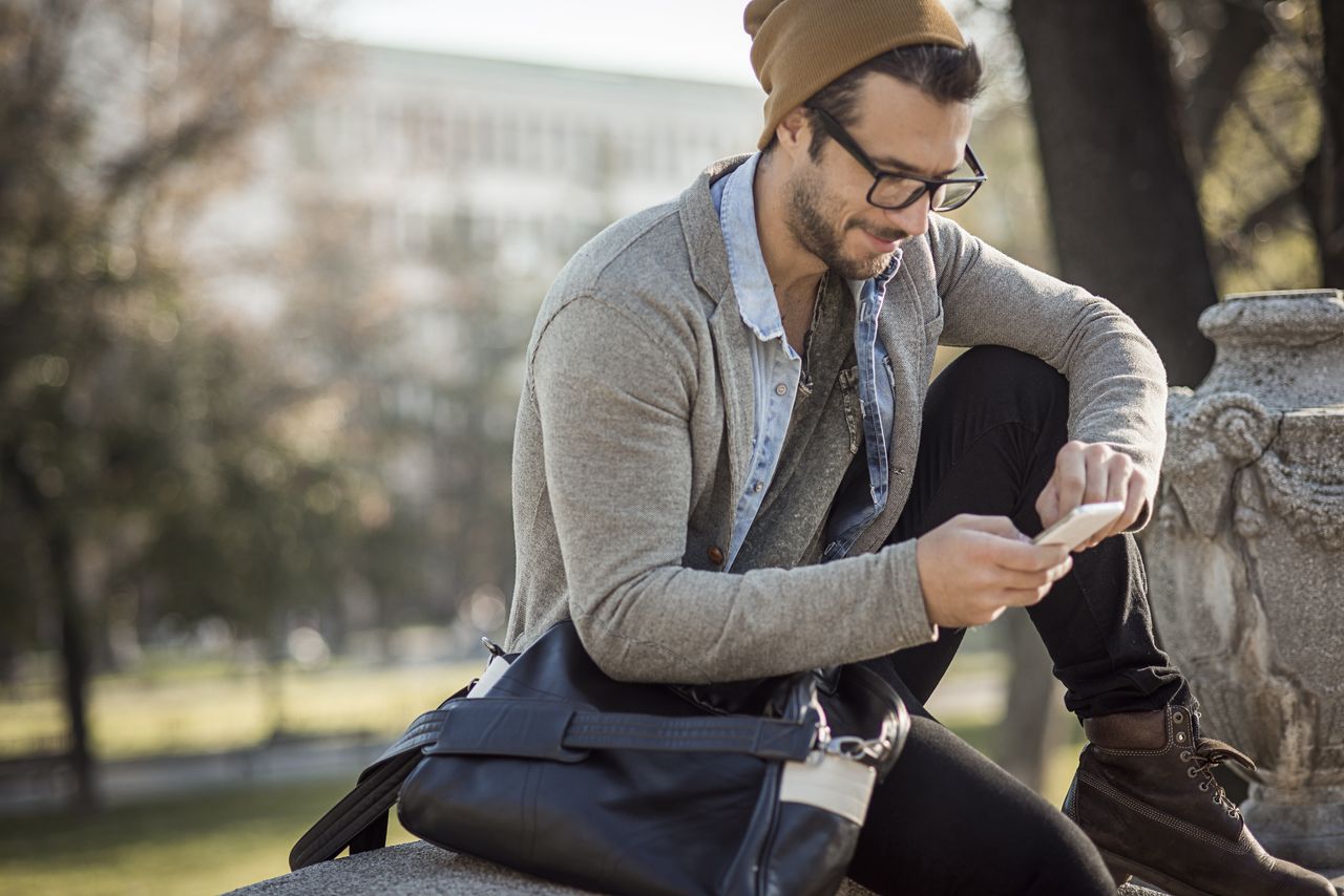 Ein casual gekleideter junger Mann mit moderner Brille und khakifarbener Mütze sitzt neben einer Steinskulptur in einem Park und lächelt beim Internetsurfen auf seinem Smartphone.