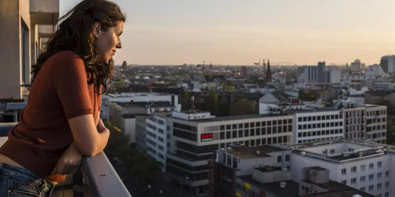 Eine junge Frau steht auf ihrem Balkon angelehnt an die Mauer und schaut auf die Stadt Berlin.
