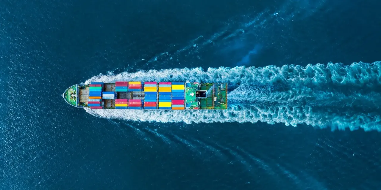 Luftaufnahme eines Containerschiffs mit bunten Frachtcontainern, das auf tiefblauem Meer fährt und eine breite Bugwelle hinterlässt.