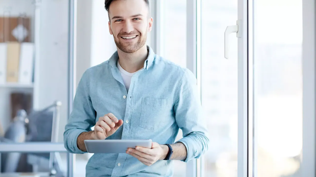 Ein braunhaariger junger Mann in einem weißen T-Shirt und einem hellblauen Jeanshemd hält seinen Tablet in einem sonnigen Büro stehend und lächelt in die Kamera.
