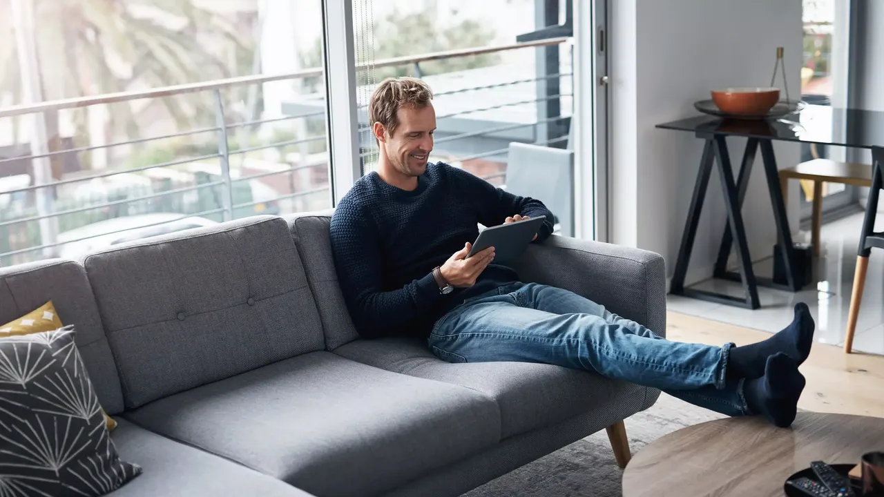 Der Mann im dunkelblauen Strickpullover sitzt auf einem grauen Sofa in einem modern ausgestatteten Zimmer mit Glaswänden und schaut lachend auf sein Tablet.