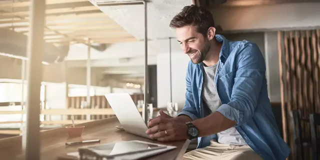 Ein junger dunkelhaariger bärtiger Mann sitzt auf dem zweiten Stock des sonnigen Open-Space-Büros in einem weißen T-Shirt, einem Denim-Hemd und in einer khakifarbigen Hose und arbeitet am Laptop, welches auf dem Holztisch neben einem Tablet und Dokumenten sich befindet.