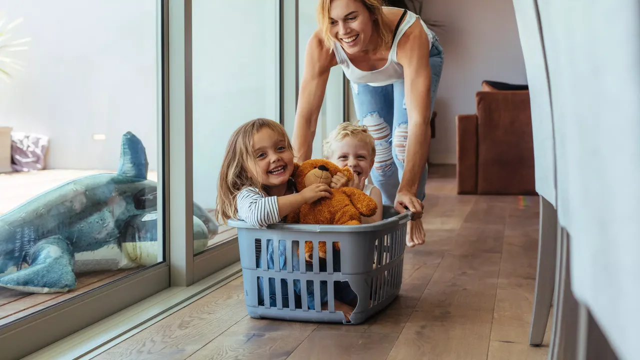 Die lächelnde Mutter mit blonden Haaren in einem weißen T-Shirt und in den Jeans spielt zu Hause mit ihren kleinen Kindern – einem blonden Mädchen mit dem braunen Teddy-Bär und einem blonden Junge, – welche in einem Wäschekorb sitzen und lächeln.