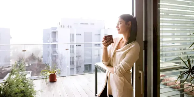 Eine junge dunkelhaarige Frau in einem beigen Cardigan steht auf der Balkonterrasse ihrer Wohnung mit einer roten Tasse mit Kaffee und blickt lächelnd in die Ferne; im Fotohintergrund ist eine Hauspflanze in einem braunen Topf sowie ein Weihnachtsbaum zu sehen.