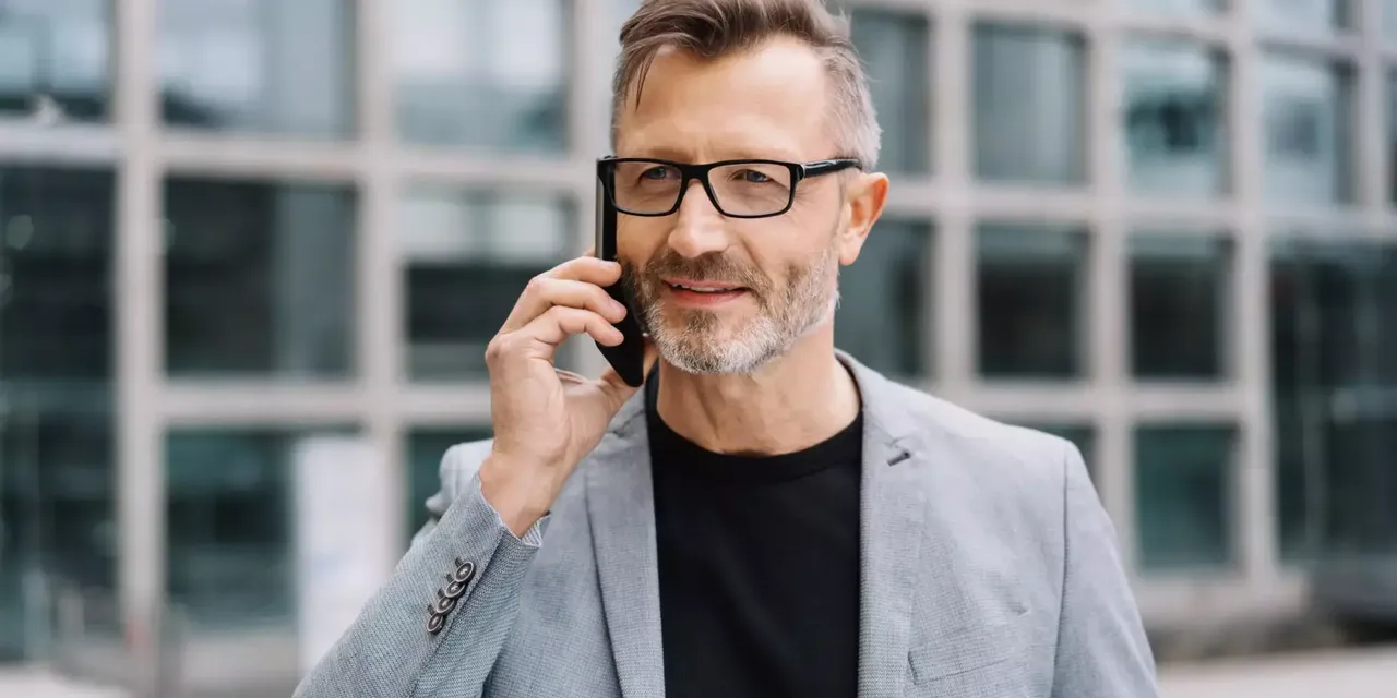 Ein Mann mittleren Alters in schwarzer Brille steht in einem hellen Anzug und einem schwarzen T-Shirt draußen vor seinem Office und telefoniert mit jemanden am Smartphone.