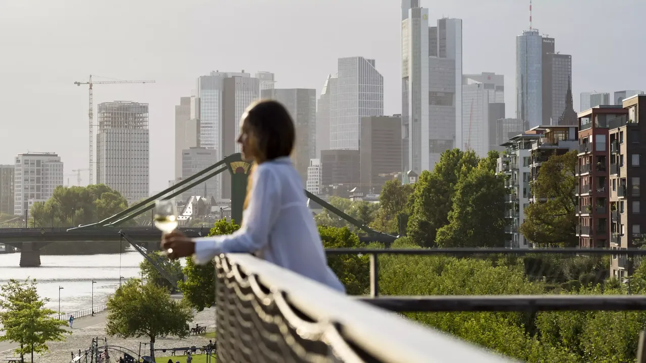 Außenansicht: Eine Frau steht auf einem Balkon der hausInvest Immobilie One Forty West in Frankfurt am Main und schaut auf die Frankfutert Skyline