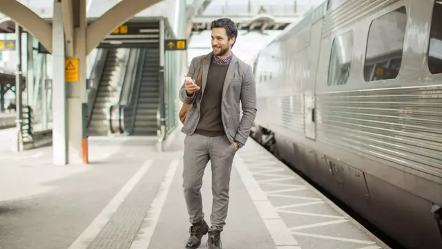 Ein bärtiger Mann in einem grauen Anzug, einem karierten Hemd und einem braunen Sweatshirt steht mit einer hellbraunen Tasche auf der rechten Schulter auf dem Bahnsteig neben einem metallgrauen Zug und tippt lächelnd auf seinem Smartphone.