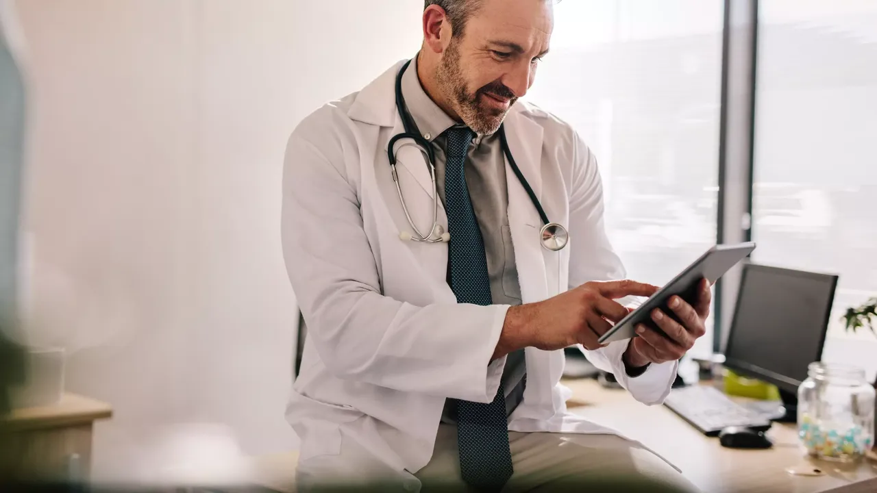Es wird ein Arzt in seiner Praxis mit einem Tablet PC in der Hand dargestellt.