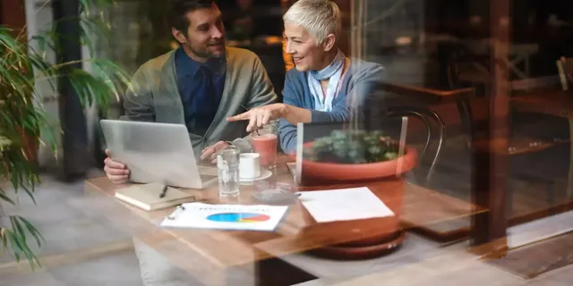 Ein dunkelhaariger Mann in einem dunkelblauen Hemd und einem grauen Cardigan richtet sein Blick auf die Blondine in einem hell-blauen Cardigan, welche auf den Bildschirm mit dem Finger zeigt; die beiden sitzen im Restaurant am Holztisch vor zwei Laptops und dem farbigen Tortendiagramm auf dem Tisch.