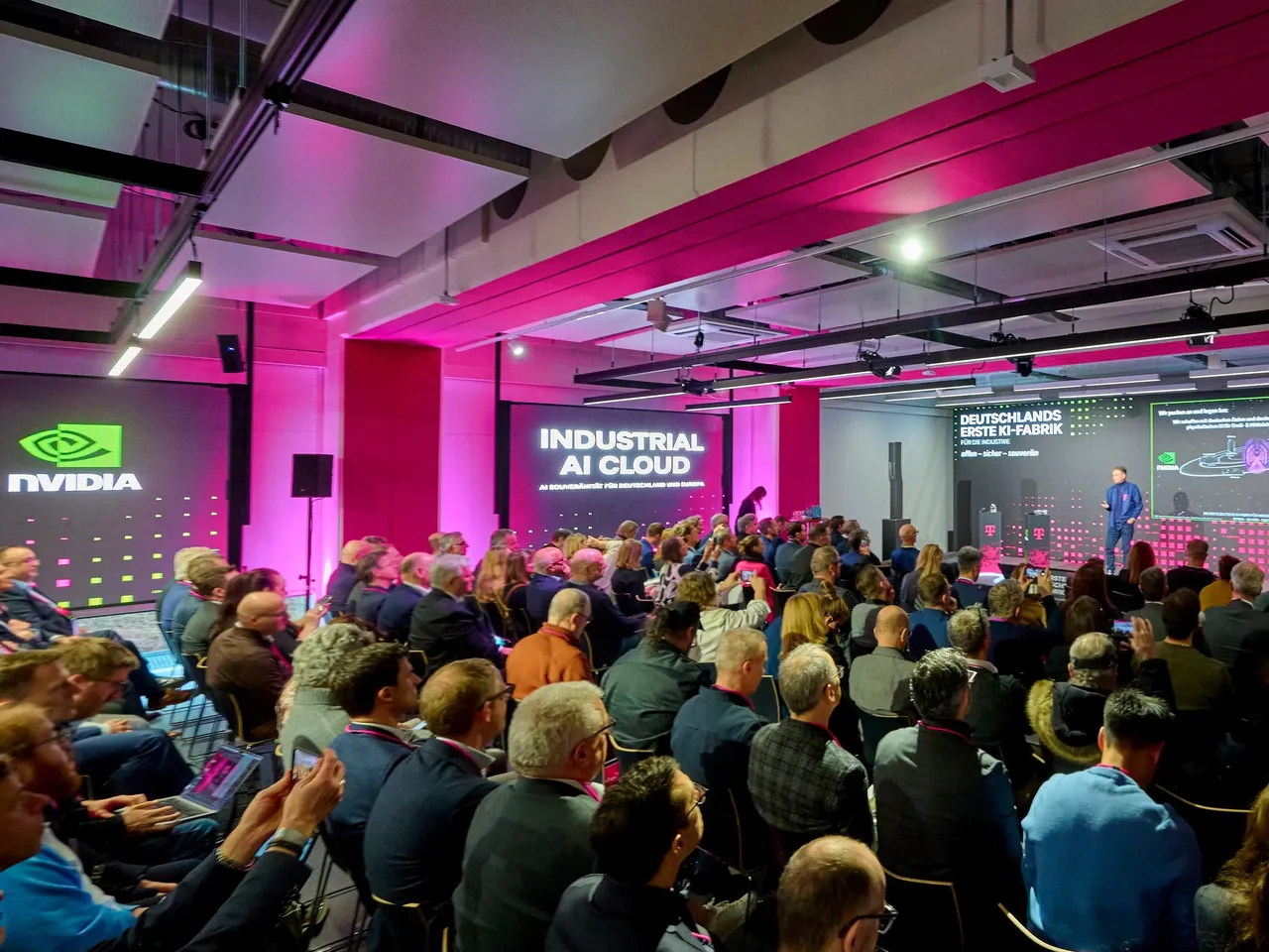 Es werden im Rahmen der Eröffnung der KI-Fabrik am Tucherpark im Eisbachviertel in München das Publikum mit zahlreichen Pressevertretern im Vordergrund dargestellt.