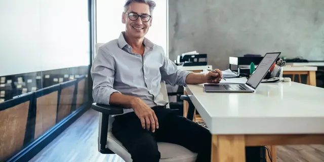 Ein Mann mittleren Alters mit schwarzer Brille sitzt in seinem Bureau am Tisch vor dem Laptop und einem kleinen künstlichen Kaktus und blickt direkt in die Kamera lächelnd.
