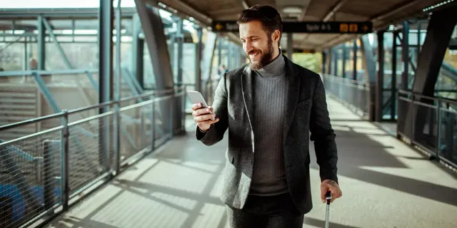 Ein dunkelhaariger bärtiger Mann in einem grauen Pullover und einer grauen Jacke steht mit seinem Koffer auf dem Bahnsteig und schaut auf sein weißfarbiges Handy.