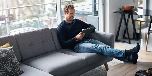 Der Mann im dunkelblauen Strickpullover sitzt auf einem grauen Sofa in einem modern ausgestatteten Zimmer mit Glaswänden und schaut lachend auf sein Tablet.