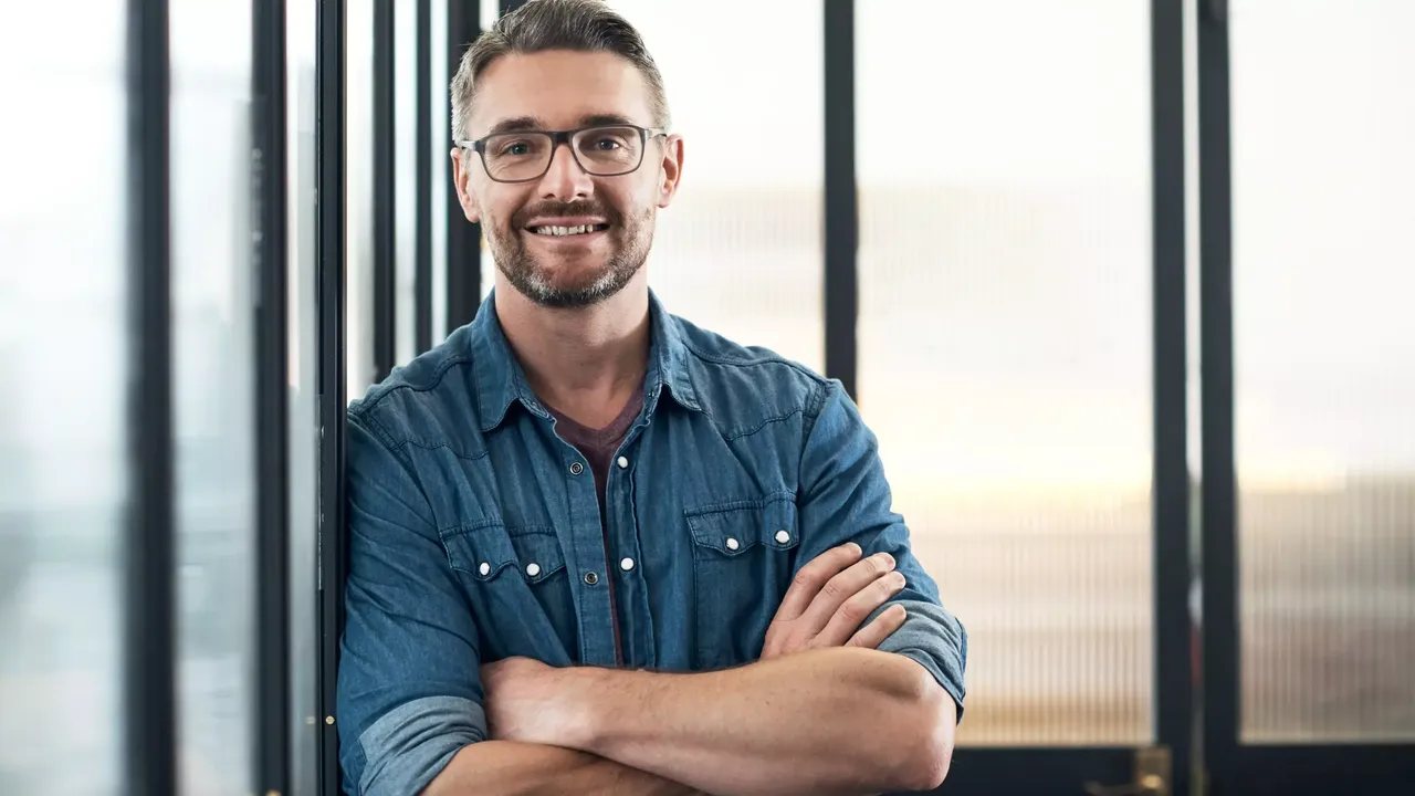 Junger Mann mit Brille und blauem Jeanshemd steht im Büro angelehnt an eine Scheibe und hat die Arme verschränkt und schaut direkt in die Kamera