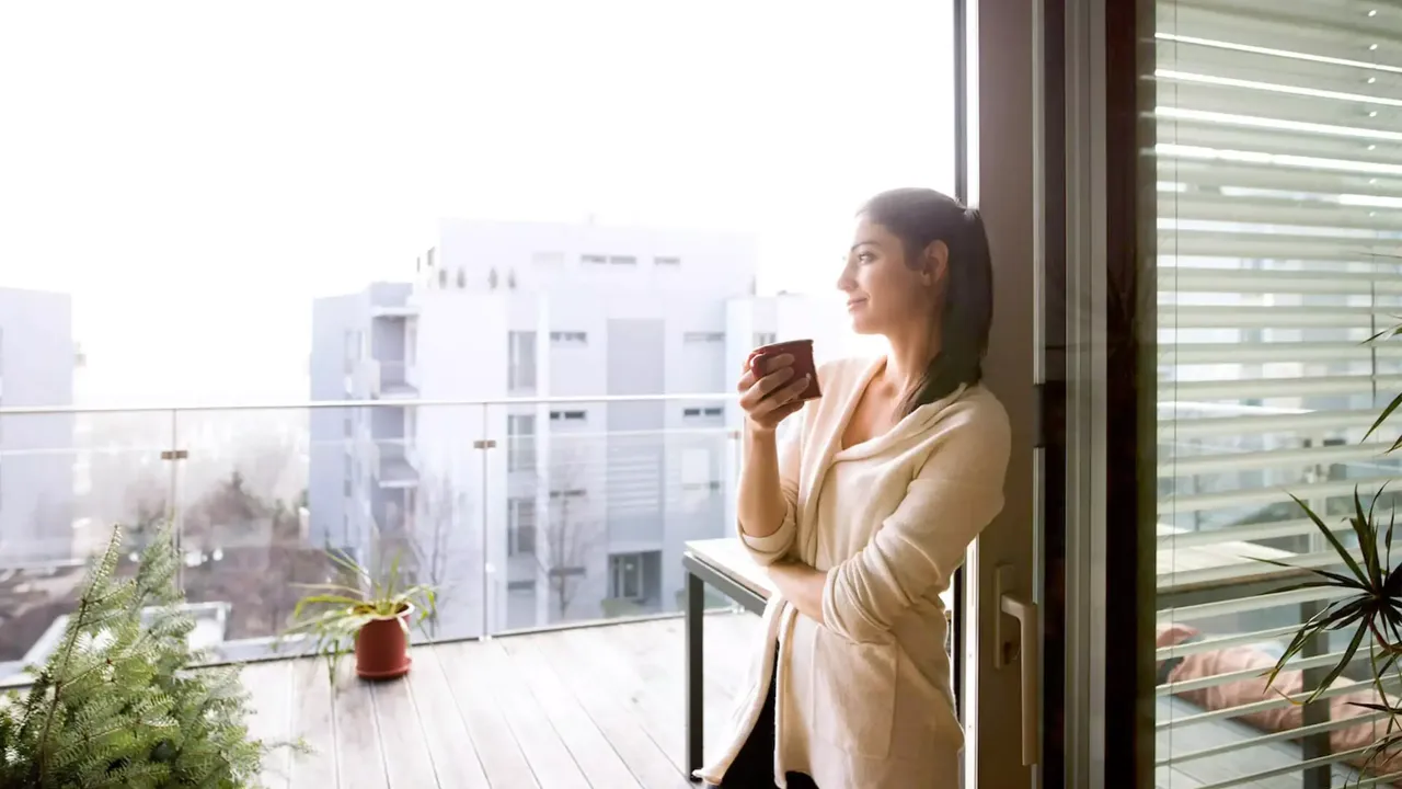 Eine junge dunkelhaarige Frau in einem beigen Cardigan steht auf der Balkonterrasse ihrer Wohnung mit einer roten Tasse mit Kaffee und blickt lächelnd in die Ferne; im Fotohintergrund ist eine Hauspflanze in einem braunen Topf sowie ein Weihnachtsbaum zu sehen.