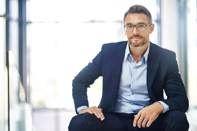 Ein bärtiger Mann mittleren Alters mit schwarzer Brille, einem dunkelblauen Anzug und einem hellbauen Hemd sitzt im Treppenhaus eines Büros mit Glaswänden und guckt direkt in die Kamera.