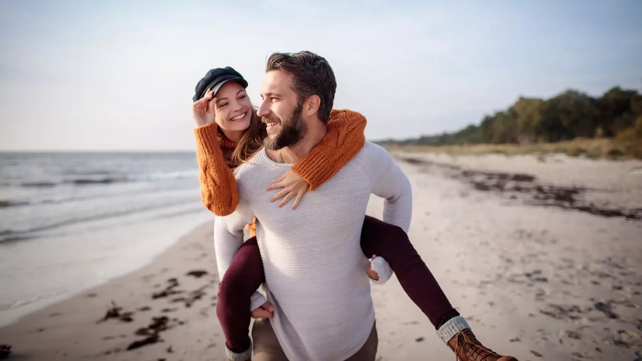 Ein Mann mittleren Alters in einem weißen Hemd trägt seine lächelnde Freundin in einem orangefarbenen Pullover und einer schwarzen Baskenmütze auf dem Rücken, während er lächelnd am Strand spazieren geht.