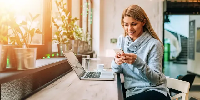 Eine Frau mit hellen Haaren sitzt lächelnd in einem grauen Kapuzenpulli auf dem Stuhl in einem Open-Space-Office mit ihrem Laptop auf dem Tisch und einer Tasse Kaffee und einer Tasse Wasser, und schreibt auf ihrem Handy.