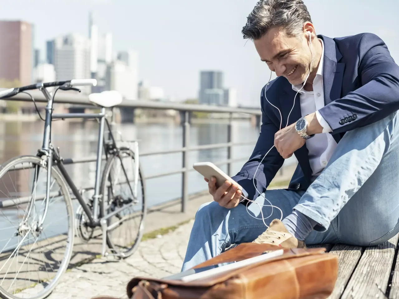 Ein Mann Mitte 40 sitzt am Mainufer in Frankfurt auf einer Holzbank und schaut auf sein Handy, neben ihm steht sein Fahrrad und seine braune Ledertasche.