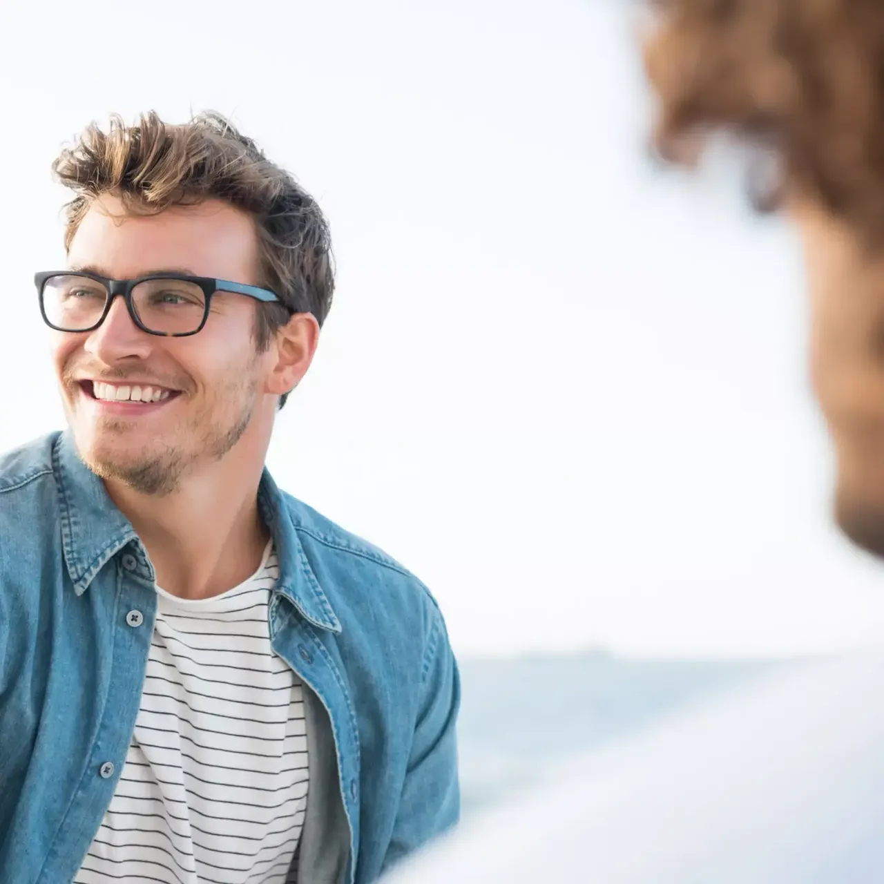 Ein junger Mann mit einem hellblauen Heansjemd und einer schwarzen Brille steht draußen und schaut lächelnd in die Kamera