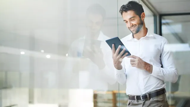 Ein junger Mann mit einem weißen Hemd steht im Büro an einer Glaswand und schaut interessiert auf sein iPad