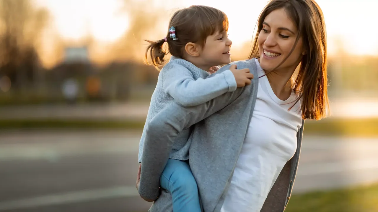 Die Mutter in einem weißen T-Shirt auf der Straße sieht ihre kleine Tochter in einem hellblauen Cardigan an, welche auf ihrem Rücken sitzt und lächelt.